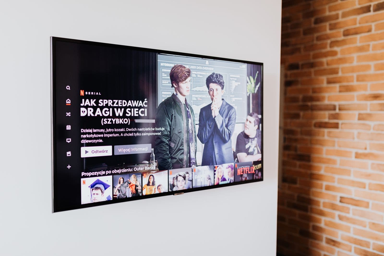A slim television mounted flush against a white wall beside an exposed-brick column